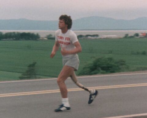 An archival image shows Terry Fox running along a rural street, wearing a white t-shirt and jogging shorts.