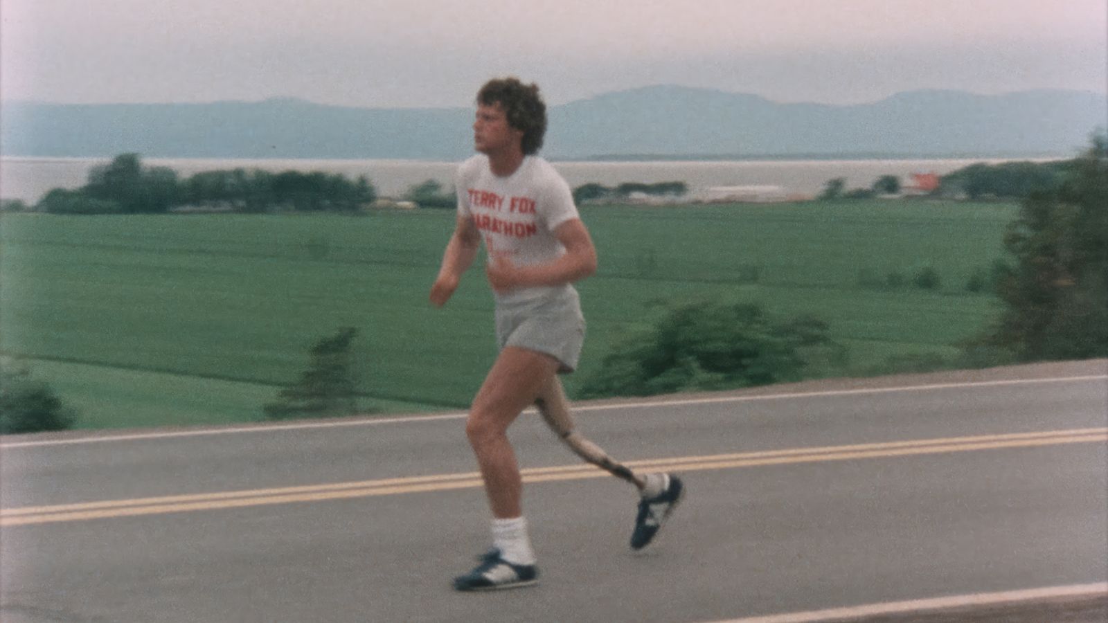 An archival image shows Terry Fox running along a rural street, wearing a white t-shirt and jogging shorts.