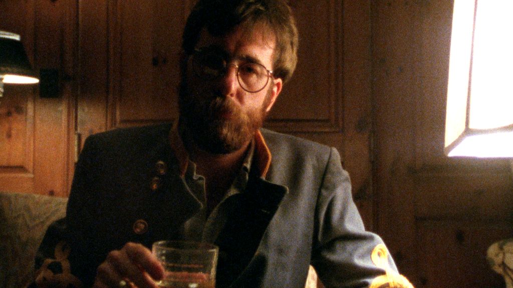 A photo shows filmmaker Ross McElwee in a scene from Sherman's March. He is sitting in a dark cabin, facing the camera, and holding a tumbler of whiskey. He is wearing an old Civil War uniform and has reddish brown hair and a bushy beard.