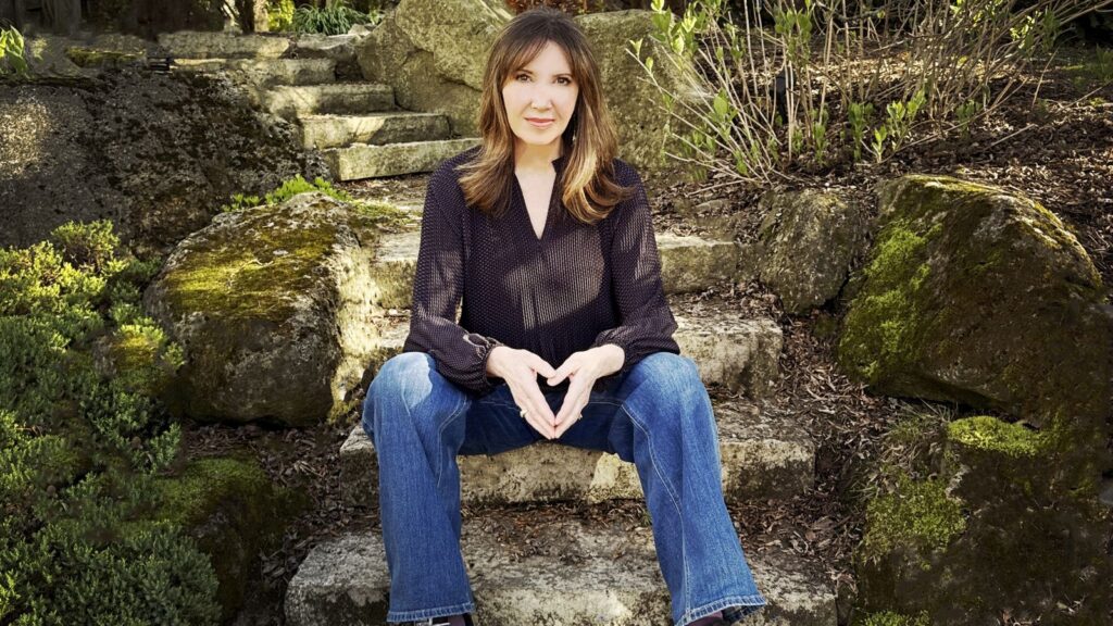 Filmmaker Shelley Saywell sits on stone steps in a garden. She is a white woman with shoulder-length blond hair. She is wearing a black shirt and blue jeans, and is looking directly at the camera.