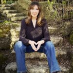 Filmmaker Shelley Saywell sits on stone steps in a garden. She is a white woman with shoulder-length blond hair. She is wearing a black shirt and blue jeans, and is looking directly at the camera.