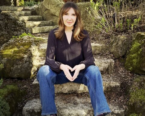 Filmmaker Shelley Saywell sits on stone steps in a garden. She is a white woman with shoulder-length blond hair. She is wearing a black shirt and blue jeans, and is looking directly at the camera.