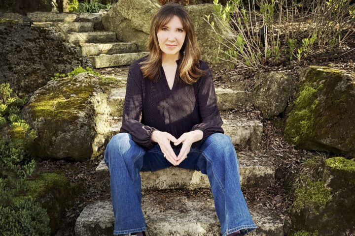 Filmmaker Shelley Saywell sits on stone steps in a garden. She is a white woman with shoulder-length blond hair. She is wearing a black shirt and blue jeans, and is looking directly at the camera.
