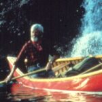 An archival image shows a man rowing a red canoe through a river.