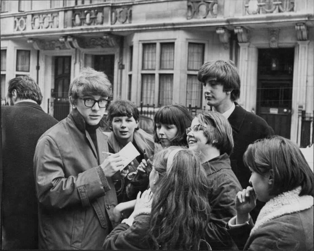 A black and white photo shows musician Peter Asher in the 1960s. He is signing autographs for a group of young women. They are on a street in London, England and all wearing overcoats. Asher is looking at the camera and wearing black glasses.