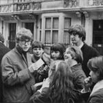A black and white photo shows musician Peter Asher in the 1960s. He is signing autographs for a group of young women. They are on a street in London, England and all wearing overcoats. Asher is looking at the camera and wearing black glasses.
