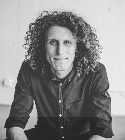 Filmmaker Andrew Morgan is pictured in a black and white portrait, shot from the waist up. He is a white man with curly shoulder-length hair and is wearing a black collared shirt