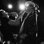 Two bluegrass singers - one woman, one man - perform face each other on stage. The photo is in black and white with dramatic lighting as spotlights appear above and behind them.