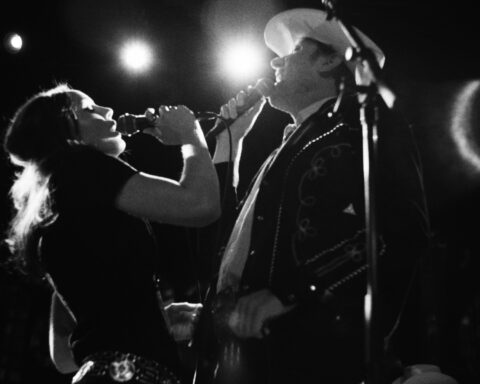 Two bluegrass singers - one woman, one man - perform face each other on stage. The photo is in black and white with dramatic lighting as spotlights appear above and behind them.