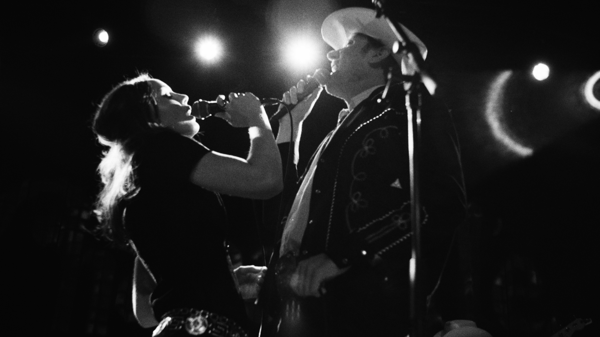 Two bluegrass singers - one woman, one man - perform face each other on stage. The photo is in black and white with dramatic lighting as spotlights appear above and behind them.