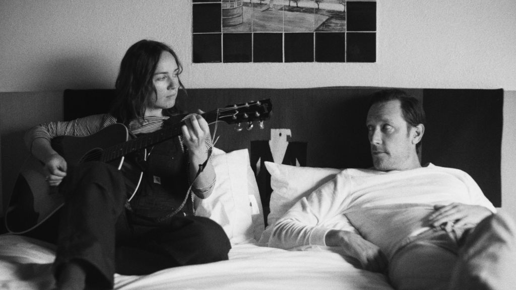 A black and  white image of a woman and  man playing guitars in bed.