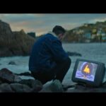 A man crouches down on a rocky shore in Newfoundland, looking at a TV that is positioned on the ground by the water. There is a strange image on the screen with a figure in a dark purple hood and an orange light where his face should be.