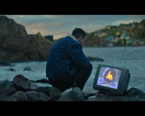 A man crouches down on a rocky shore in Newfoundland, looking at a TV that is positioned on the ground by the water. There is a strange image on the screen with a figure in a dark purple hood and an orange light where his face should be.