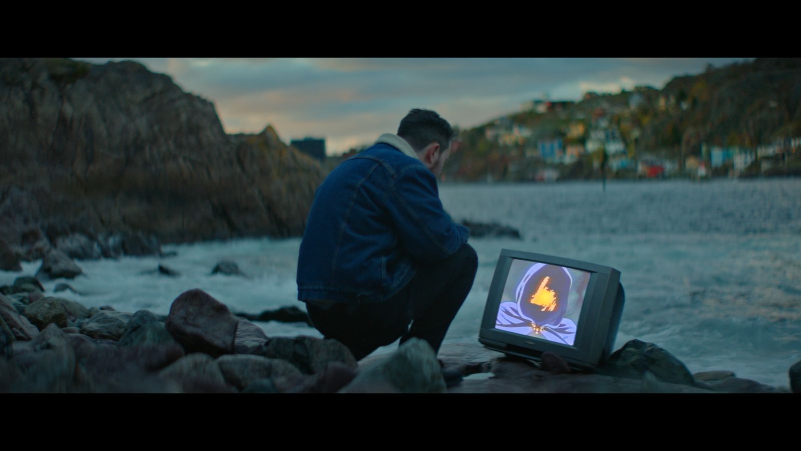 A man crouches down on a rocky shore in Newfoundland, looking at a TV that is positioned on the ground by the water. There is a strange image on the screen with a figure in a dark purple hood and an orange light where his face should be.