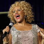Darlene Love appears on stage performing a song. She is Black woman with blonde curly hair. She is holding a microphone and leaning back, and wearing a silver sequined dress.