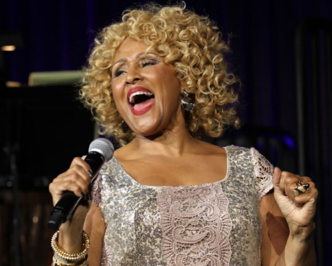Darlene Love appears on stage performing a song. She is Black woman with blonde curly hair. She is holding a microphone and leaning back, and wearing a silver sequined dress.