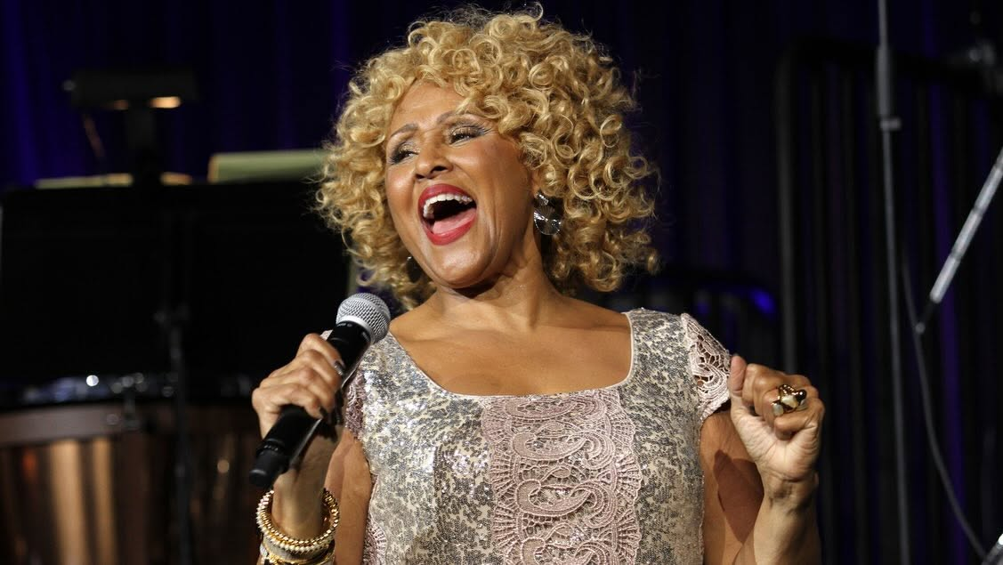 Darlene Love appears on stage performing a song. She is Black woman with blonde curly hair. She is holding a microphone and leaning back, and wearing a silver sequined dress.