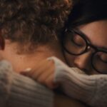 A Filipina woman with black glasses wmbraces her white partner. They are pictured in close-up.