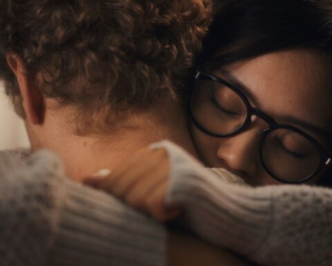A Filipina woman with black glasses wmbraces her white partner. They are pictured in close-up.