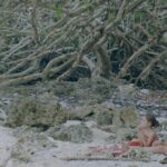 A wide shot shows a mother and her child sitting on a beach. There are many branches behind them.