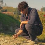 A young Italian man with brown curly hair crouches over a dry patch of earth in a vineyard, holding a handful of white clay.