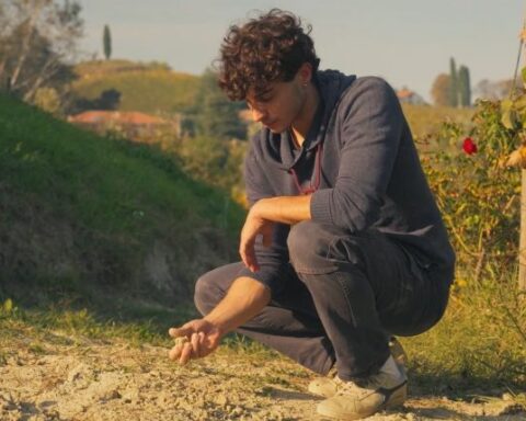 A young Italian man with brown curly hair crouches over a dry patch of earth in a vineyard, holding a handful of white clay.