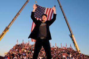 Elon Musk stands before a crowed in a stadium. He has both hands raised in the air, and there is an American flag behind him. He is wearing a black jacket and baseball cap.