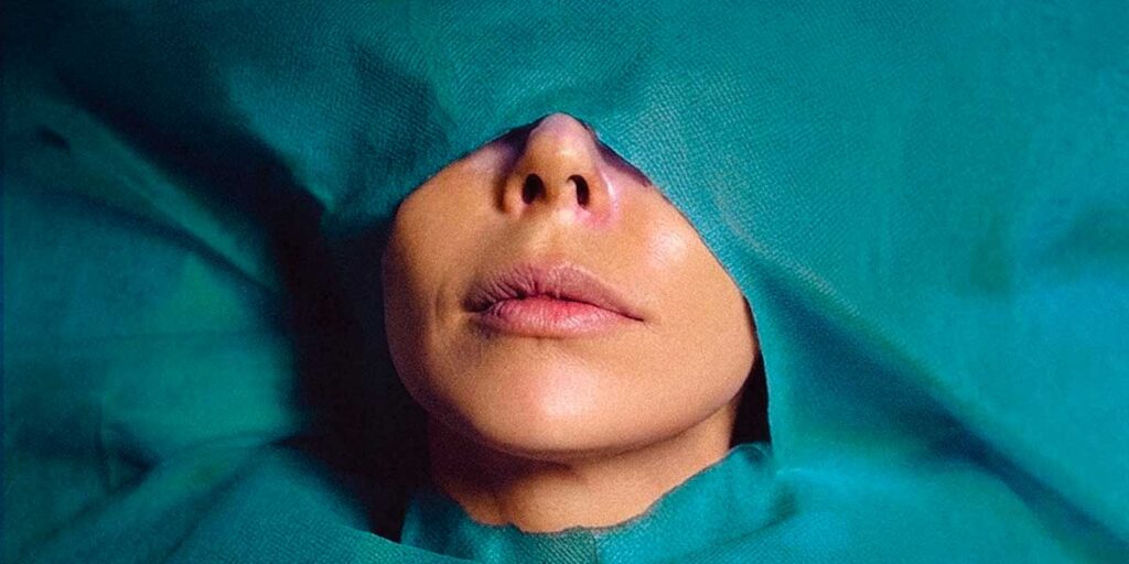 An Iranian woman is pictured in close up, lying beneath a teal surgical sheet. The sheet covers her body, with a hole cut that exposes her face from the nose to her chin.