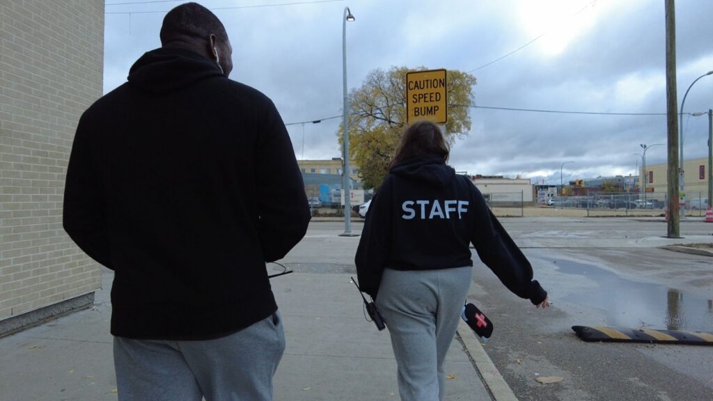 A view from behind of a tall man and shorter woman as they walk through a city street. They are both wearing black sweatshirts. The woman's shirt says STAFF in white letters.