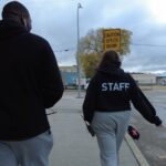 A view from behind of a tall man and shorter woman as they walk through a city street. They are both wearing black sweatshirts. The woman's shirt says STAFF in white letters.