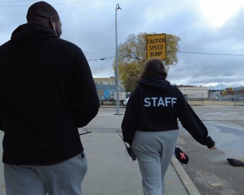 A view from behind of a tall man and shorter woman as they walk through a city street. They are both wearing black sweatshirts. The woman's shirt says STAFF in white letters.