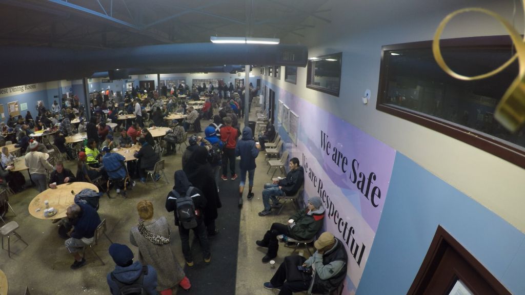 A high angle view of a community shelter. Lots of people are seated at tables. The words 'We Are Safe' appear in large purple letters on a white wall.