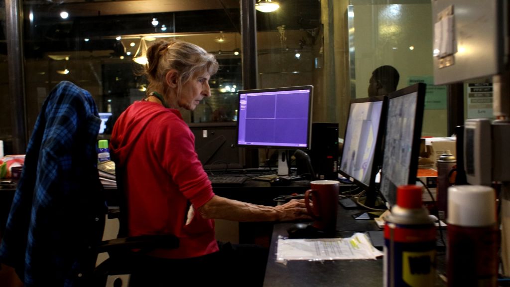 A woman sits at a desk with two computer monitors. She is a white woman with blond hair and is wearing a red sweatshirt.