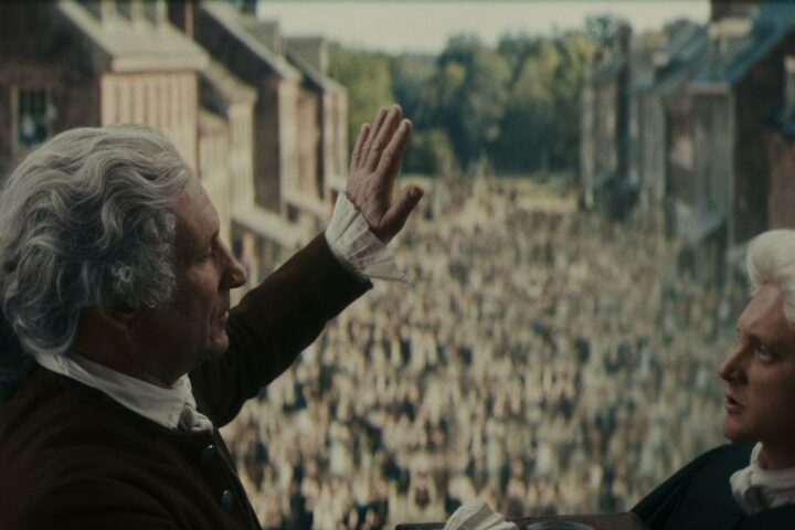 Two men in Civil War era America speak before a crowd of people. The men have white wigs and black robes.