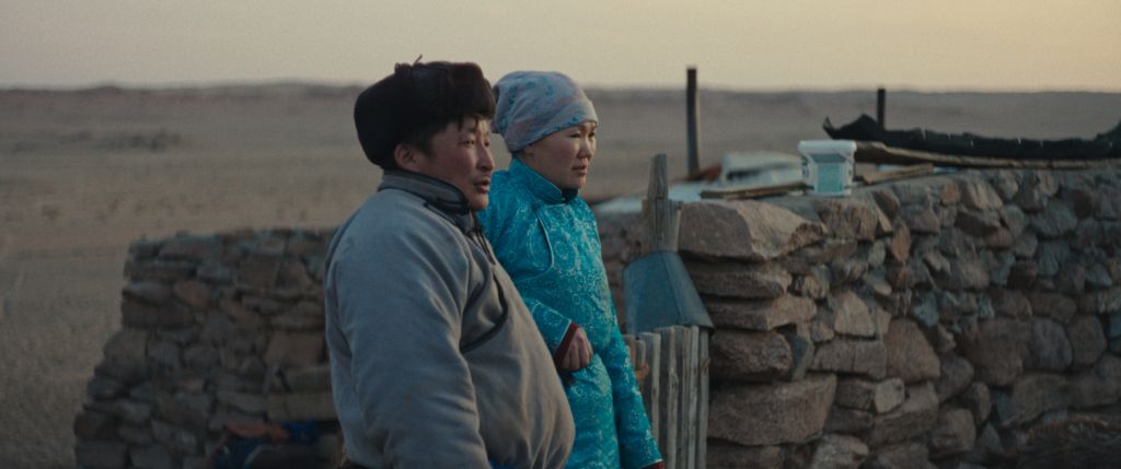 Two Mongolian farmers (a husband and wife) stand outside by a stone wall, observing something in the distance.