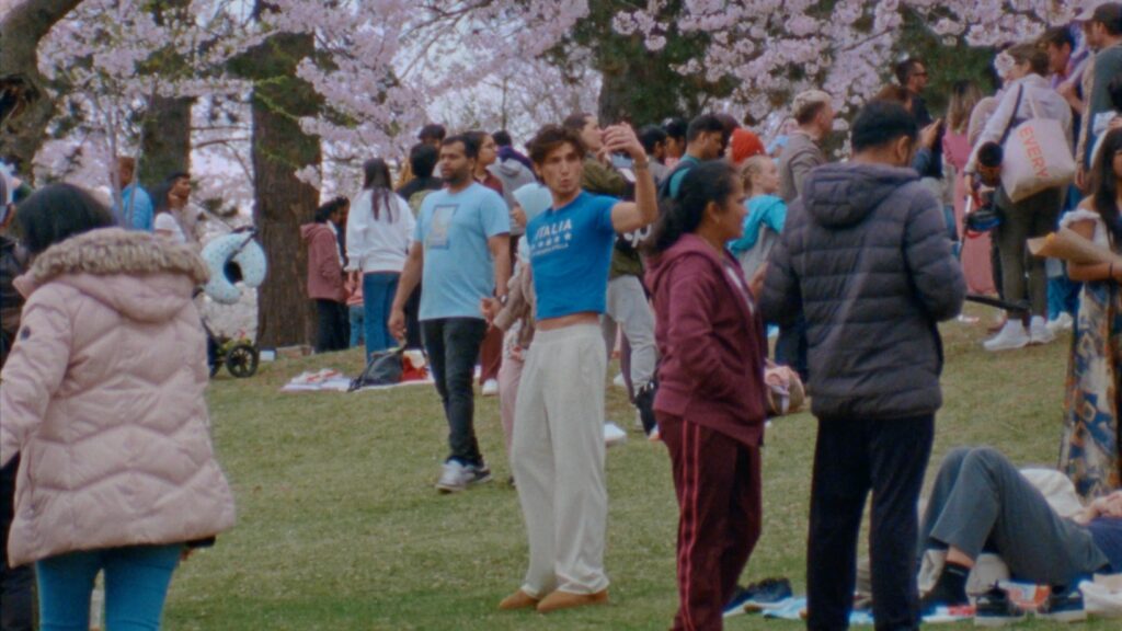 An influencer takes a selfie in High Park with many cherry blossoms in the background. He is a skinny white boy in a boy cropped t-shirt and white pants. The park is busy and full of pink cherry blossoms on the trees.