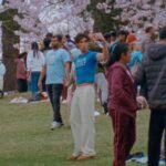 An influencer takes a selfie in High Park with many cherry blossoms in the background. He is a skinny white boy in a boy cropped t-shirt and white pants. The park is busy and full of pink cherry blossoms on the trees.