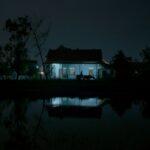 A dark photo depicts the silhouette of a man as he walks outside a home. The house has a light on and casts a blue glow.