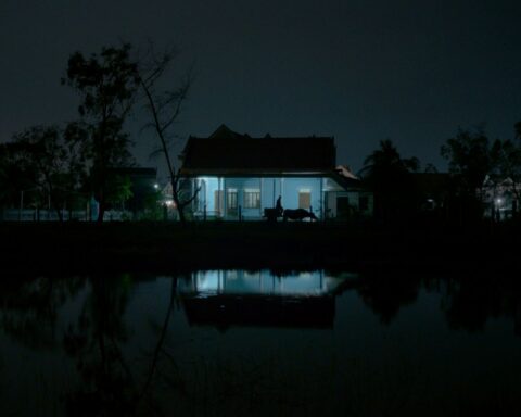 A dark photo depicts the silhouette of a man as he walks outside a home. The house has a light on and casts a blue glow.