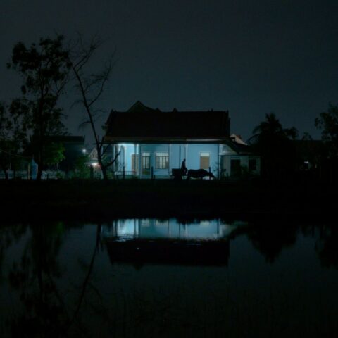 A dark photo depicts the silhouette of a man as he walks outside a home. The house has a light on and casts a blue glow.