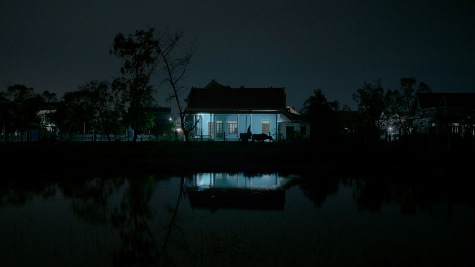 A dark photo depicts the silhouette of a man as he walks outside a home. The house has a light on and casts a blue glow.