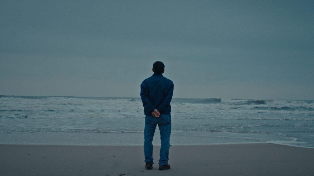 A Vietnamese man stands on a beach at the edge of the water, looking out towards the horizon. His back is to the camera and the sky is overcast.