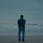 A Vietnamese man stands on a beach at the edge of the water, looking out towards the horizon. His back is to the camera and the sky is overcast.