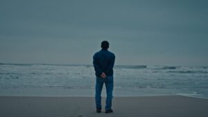 A Vietnamese man stands on a beach at the edge of the water, looking out towards the horizon. His back is to the camera and the sky is overcast.