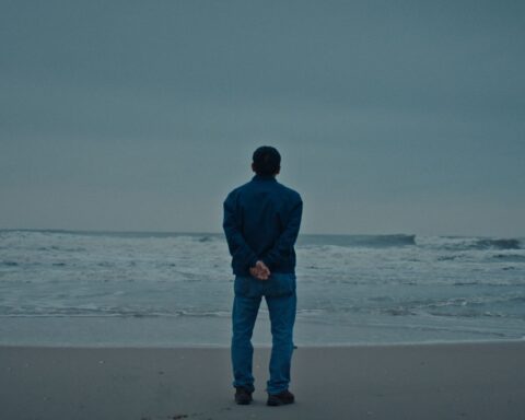 A Vietnamese man stands on a beach at the edge of the water, looking out towards the horizon. His back is to the camera and the sky is overcast.
