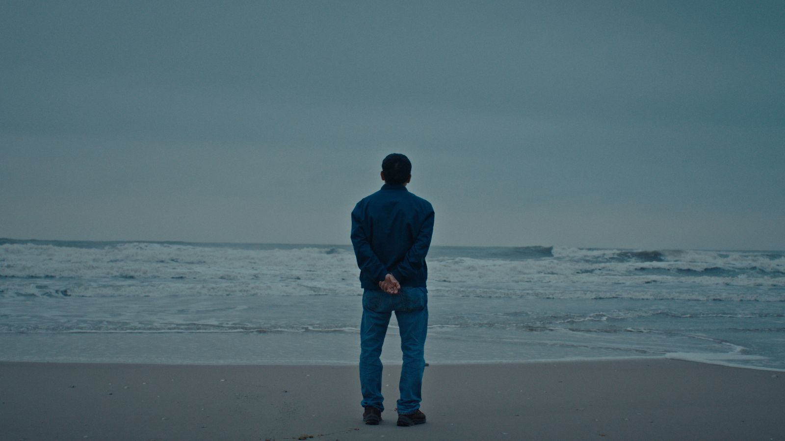 A Vietnamese man stands on a beach at the edge of the water, looking out towards the horizon. His back is to the camera and the sky is overcast.