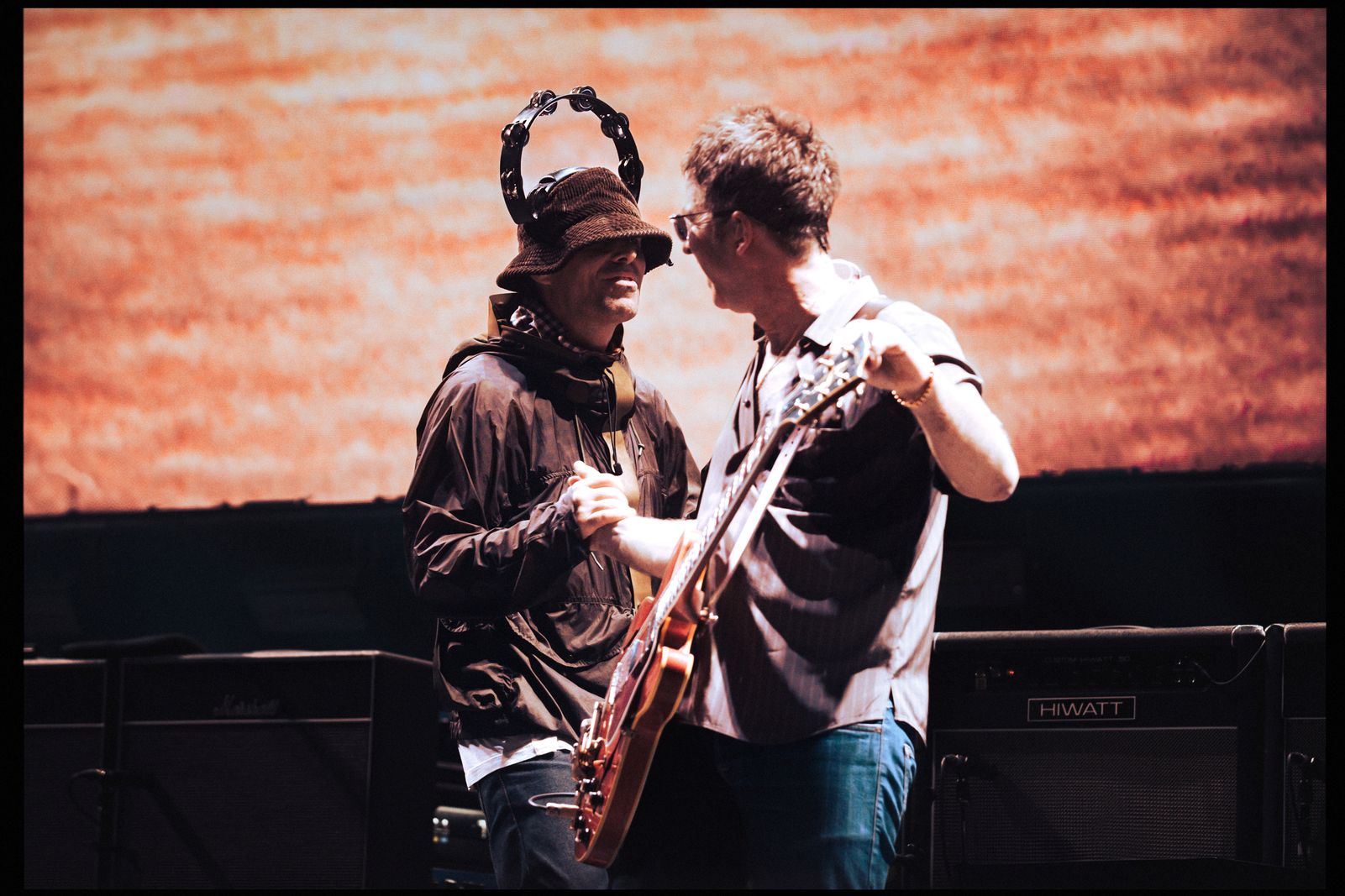 Two middle-aged male rock stars shake hands on stage. They are both white men holding guitars. The one on the left is wearing a brown jacket and brown bucket hat, and has a tambourine on his head. The man on the right is wearing blue jeans and a grey collared shirt. The large screen behind them has a red pattern.