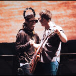 Two middle-aged male rock stars shake hands on stage. They are both white men holding guitars. The one on the left is wearing a brown jacket and brown bucket hat, and has a tambourine on his head. The man on the right is wearing blue jeans and a grey collared shirt. The large screen behind them has a red pattern.