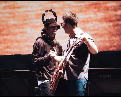 Two middle-aged male rock stars shake hands on stage. They are both white men holding guitars. The one on the left is wearing a brown jacket and brown bucket hat, and has a tambourine on his head. The man on the right is wearing blue jeans and a grey collared shirt. The large screen behind them has a red pattern.