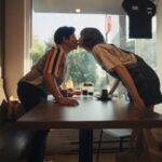 Two women--one Filipina, one white--stand up and lean over a table to share a kiss.